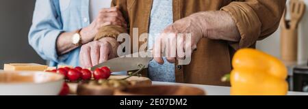 vista ritagliata di una donna anziana vicino all'uomo tagliando il formaggio sul tavolo con verdure in primo piano sfocato, banner Foto Stock