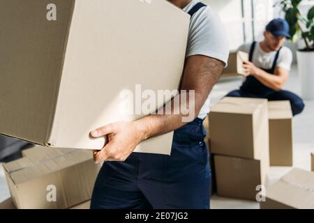 vista ritagliata del mover indiano che tiene la scatola di cartone vicino al coworker su sfondo sfocato Foto Stock