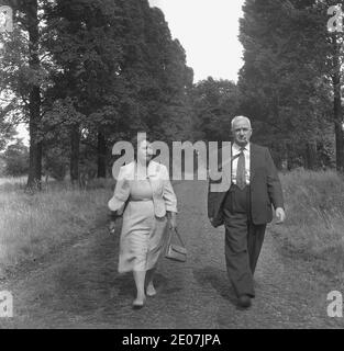 Anni '50, storica, una coppia di mezza età, ben vestito, che cammina lungo un percorso erboso alberato in un parco semi-rurale, Inghilterra, Regno Unito. Il Signore indossa un vestito e cravatta e fuma un tubo, mentre la signora sta portando la sua borsa, forse stanno facendo una passeggiata dopo aver frequentato la chiesa o una funzione civica. Foto Stock
