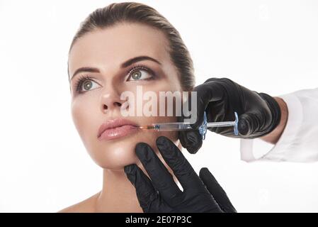 Concetto di aumento delle labbra. Mani in guanti neri che fanno l'iniezione di riempitivo alle labbra attraenti della donna. Ritratto su sfondo bianco. Foto Stock