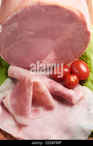 Delizioso prosciutto cotto a fette e arrotolato su tavola di legno accompagnato da pomodori ciliegini freschi e lattuga. Ampia vista con un grande pezzo di prosciutto, vista dall'alto Foto Stock