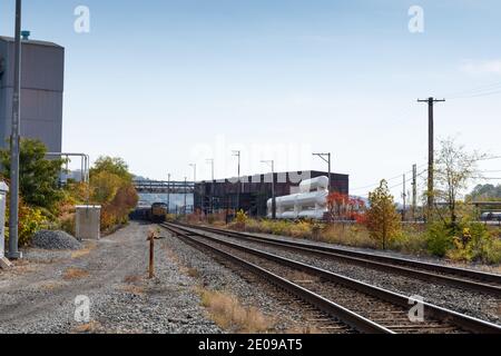 Treno che scende su una pista che corre lungo edifici industriali in un'area rurale, stagione autunnale con foglie autunnali, aspetto orizzontale Foto Stock