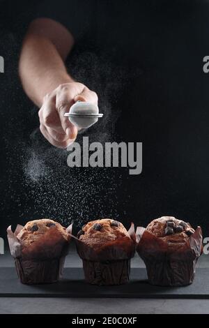 Mani che versano zucchero in polvere su muffin al cioccolato su sfondo nero. Formato verticale. Foto Stock