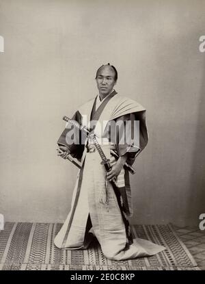 fotografia d'epoca del xix secolo - samurai con due spade vestite di pantaloni lunghi, ambiente studio. Giappone, circa 1880. Probabilmente posto da un attore. Foto Stock