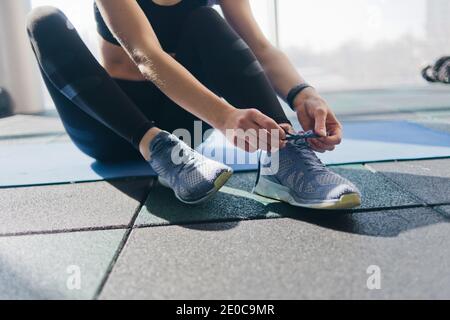 Donna che lega i lacci delle sneakers di sport sedendosi su un tappetino in palestra Foto Stock