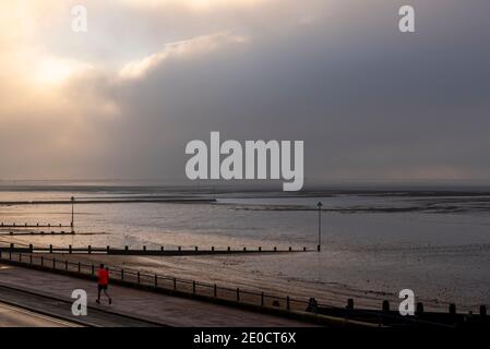 Jogger in giacca di colore brillante che corre lungo la passeggiata lungomare a Southend on Sea, Essex, Regno Unito, con il fronte meteo nero in movimento. Previsione neve Foto Stock