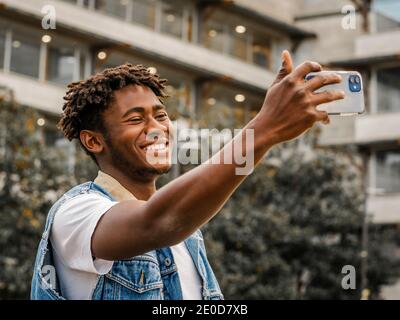 Vista laterale del giovane allegro ragazzo hipster afroamericano con i dreadlock che prendono selfie sul telefono cellulare per la condivisione con amici nel social netw Foto Stock