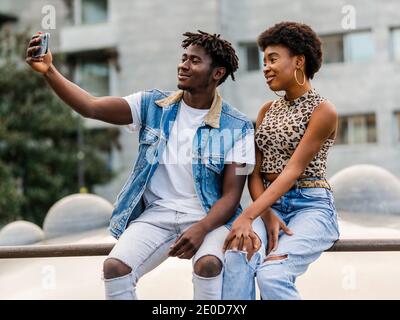 Allegro giovane afroamericano uomo e donna con capelli ricci vestito in jeans strappati alla moda seduta su recinto e prendendo selfie su smartphone, mentre sp Foto Stock