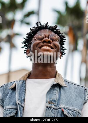 Basso angolo di felice giovane afroamericano maschio con dreadlock indossa una giacca in denim alla moda e occhiali che sorridono felicemente mentre sei in piedi sulla strada Foto Stock