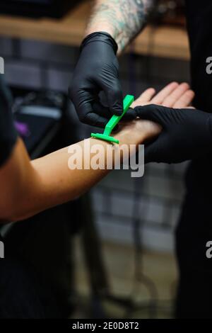 Dall'alto del raccolto anonimo maschio tatuaggio master in lattice guanti braccio di rasatura di cliente femminile irriconoscibile prima della verniciatura Foto Stock