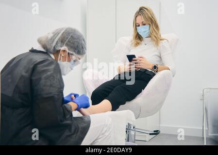 Poologo femminile in uniforme facendo pedicure per la donna seduta in sedia medica in clinica di bellezza Foto Stock