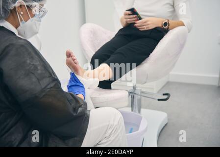 Poologo femminile in uniforme facendo pedicure per la donna seduta in sedia medica in clinica di bellezza Foto Stock