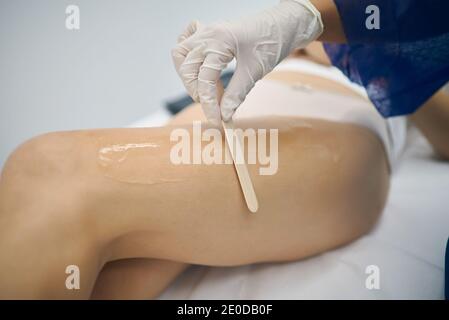 Estetista irriconoscibile che applica la pasta di zucchero con la spatola di legno sulla gamba del cliente durante la rimozione dei capelli durante la procedura di cura della pelle in clinica di bellezza Foto Stock