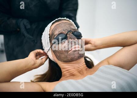 Cliente femminile con lozione di carbonio sul viso che mette su protettivo occhiali mentre si preparano per la buccia laser nel moderno centro di bellezza Foto Stock