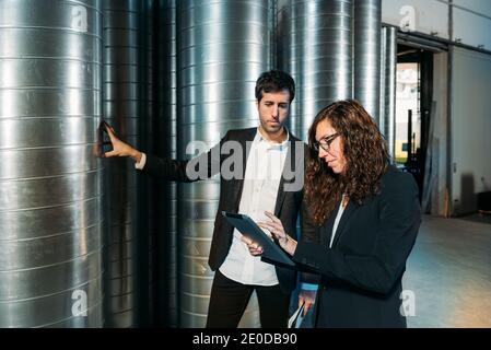 Impegnati colleghi di sesso maschile e femminile in abiti formali utilizzando tablet insieme lavorando in magazzino con barili in metallo Foto Stock