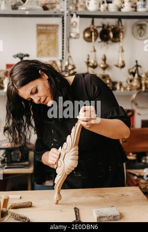 Abile donna artigianale lucidante decorativo intagliato stampaggio in legno con abrasivo carta mentre si lavora in officina professionale Foto Stock