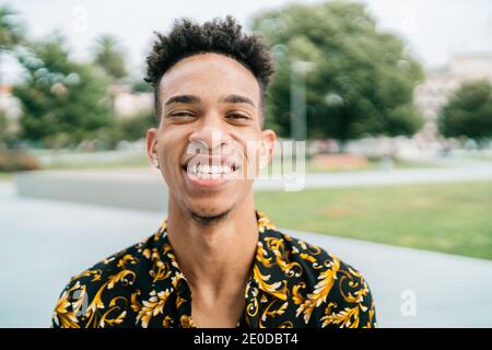Ottimista afroamericano maschio in abbigliamento fantasia e con Afro acconciatura in piedi sulla strada e sinceramente sorridente alla macchina fotografica Foto Stock