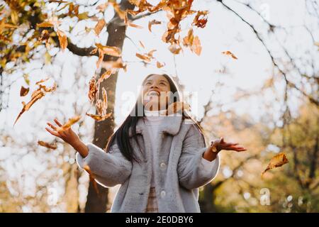 Basso angolo di femmina etnico spensierato in caldo cappotto tossing autunno caduto foglie in parco il giorno di sole mentre hanno divertimento e guardare in su Foto Stock