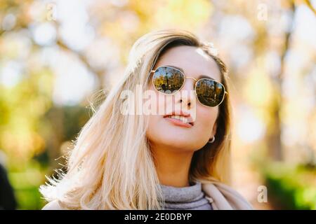 Donna sorridente che indossa cappotto autunnale e occhiali da sole in piedi nel parco in giornata di sole e guardando la fotocamera Foto Stock