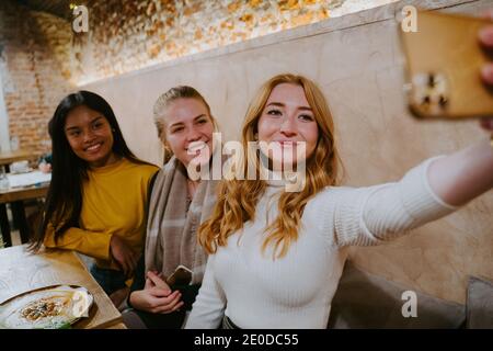 Gruppo di allegre e diverse amiche sedute al tavolo caffè e prendere selfie su smartphone mentre si passa il weekend insieme Foto Stock