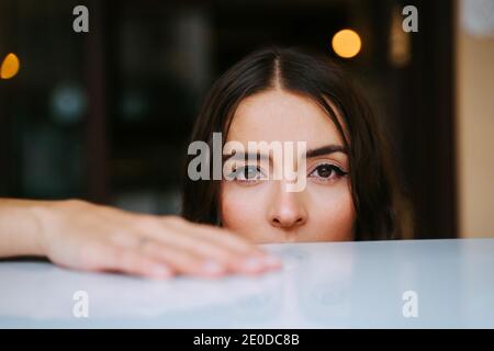 Serena giovane donna fiduciosa con lunghi capelli scuri guardando la macchina fotografica mentre riposa al tavolo nel caffè Foto Stock