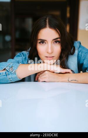 Bruna giovane calma positiva in denim e con tatuaggio sul braccio guardando la fotocamera mentre si riposa sul tavolo caffè Foto Stock