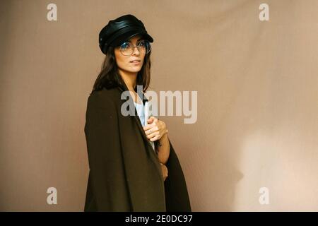 Giovane donna sicura con elegante cappuccio e pantaloni in pelle nera con cappotto trendy e occhiali che tengono carte e guardando mentre si è in piedi un Foto Stock