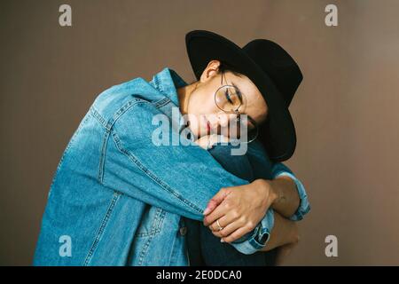 Vista laterale di una donna calmo che indossa abiti in denim seduti sfondo marrone con cappello alla moda con gli occhi chiusi e avvolgente ginocchia Foto Stock