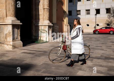 Vista laterale completa del corpo di una giovane donna elegante e calda cappotto a piedi con bicicletta retrò decorata con fiori su pavimentato strada urbana vicino al vecchio edificio Foto Stock