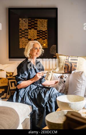 Donna matura e pensiva pacifica in elegante abito bevendo caffè e. accarezzare il cane mentre ci si rilassa su un comodo e morbido divano con cuscini in elegante soggiorno Foto Stock