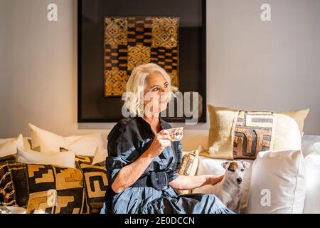 Donna matura e pensiva pacifica in elegante abito bevendo caffè e. accarezzare il cane mentre ci si rilassa su un comodo e morbido divano con cuscini in elegante soggiorno Foto Stock