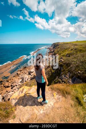 Vista posteriore di una donna irriconoscibile che si erge su una roccia ruvida strada verso il mare contro il cielo soleggiato durante le vacanze estive in Santander Foto Stock