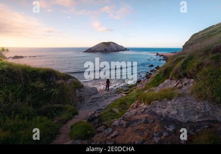 Vista posteriore di una donna irriconoscibile che cammina su una roccia ruvida strada verso il mare tempestoso contro il cielo del tramonto durante le vacanze estive A Santander Foto Stock