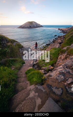 Vista posteriore di una donna irriconoscibile che cammina su una roccia ruvida strada verso il mare tempestoso contro il cielo del tramonto durante le vacanze estive A Santander Foto Stock