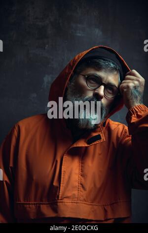 Elegante ed elegante maschio etnico adulto tatuato con barba grigia giacca trendy con cappuccio e occhiali che si affaccia su sfondo nero Foto Stock