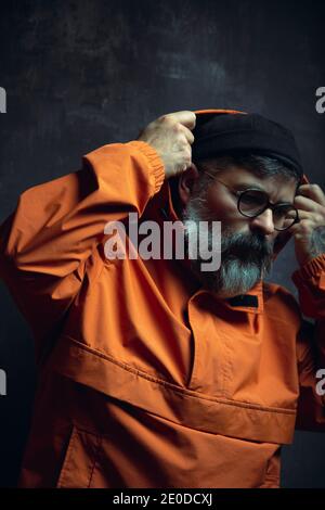 Elegante ed elegante maschio etnico adulto tatuato con barba grigia giacca trendy con cappuccio e occhiali che si affaccia su sfondo nero Foto Stock
