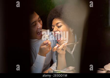 Felice uomo asiatico che alimenta allegra ragazza afroamericana con insalata mentre si diverte durante il pranzo in caffetteria Foto Stock