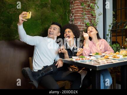 Gruppo di giovani amici multirazziali positivi che si divertono e prendono selfie su smartphone mentre si gusta un pasto delizioso insieme in un ambiente accogliente caffè Foto Stock
