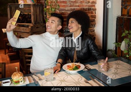 Felice giovane asiatico e ragazza afroamericana divertirsi e prendere selfie su smartphone durante il pranzo in ristorante Foto Stock