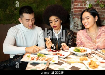 Gruppo multirazziale di giovani amici seduti a tavola con piatti insieme mentre si diverte durante la cena in un accogliente ristorante Foto Stock