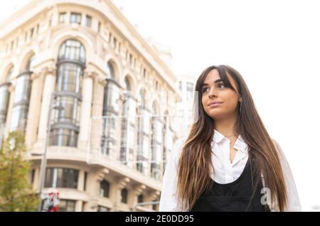 Vista laterale di una giovane donna etnica allegra con un lungo buio capelli in camicia bianca in piedi sulla strada della città vicino autobus fermati e sorridi Foto Stock