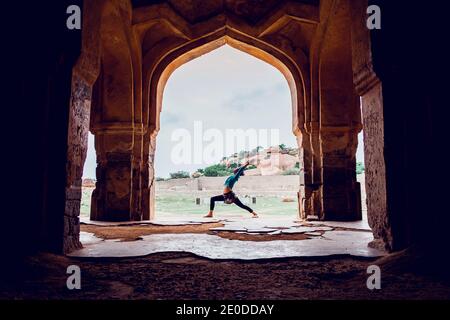 Vista laterale di una femmina non riconoscibile in Activewear Performing Warrior Poso mentre esercitavo yoga vicino al vecchio palazzo orientale arcuato in montagna te Foto Stock