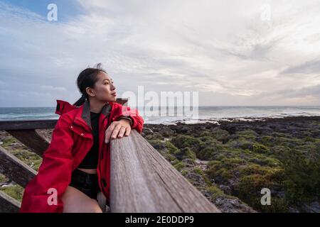 Vista laterale della giovane donna asiatica con un impermeabile rosso allontanati con attenzione mentre ti riposi da solo in riva al mare Foto Stock