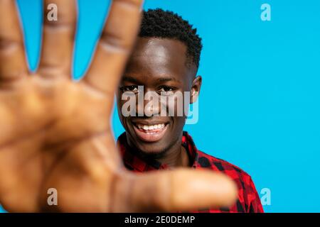 Positivo bel maschio afroamericano in camicia a scacchi rossa in piedi su sfondo blu vibrante in studio e raggiungendo la mano verso fotocamera Foto Stock