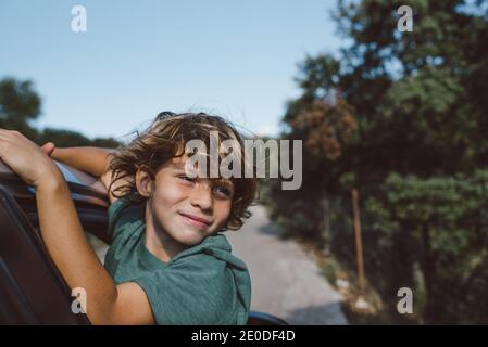 Vista laterale del ragazzo presagomato con i capelli ricci seduti dentro apri la finestra dell'automobile e guarda lontano mentre ti godi l'estate avventura in terra montuosa Foto Stock