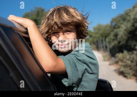 Vista laterale del ragazzo presagomato con i capelli ricci seduti dentro apri la finestra dell'automobile e guarda la fotocamera mentre ti godi avventura estiva in montagna lan Foto Stock