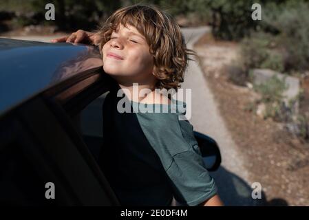 Vista laterale del ragazzo presagomato con i capelli ricci seduti dentro apri la finestra dell'automobile con gli occhi chiusi mentre ti godi l'estate avventura in terra montuosa Foto Stock