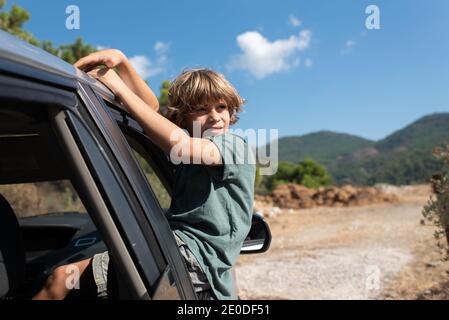 Vista laterale del ragazzo presagomato con i capelli ricci seduti dentro apri la finestra dell'automobile e guarda lontano mentre ti godi l'estate avventura in terra montuosa Foto Stock