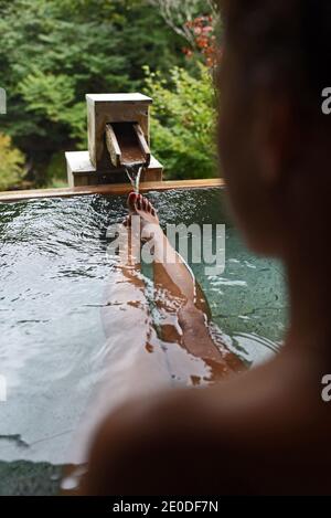 Gambe e piedi di una donna caucasica viaggiatore godendo di un bagno di acque termali in onsen giapponese, vista da dietro Foto Stock