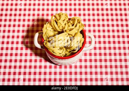 Dall'alto si trovano deliziosi nidi di pasta di fettuccine non cotti pentola su tovaglia a scacchi in cucina Foto Stock
